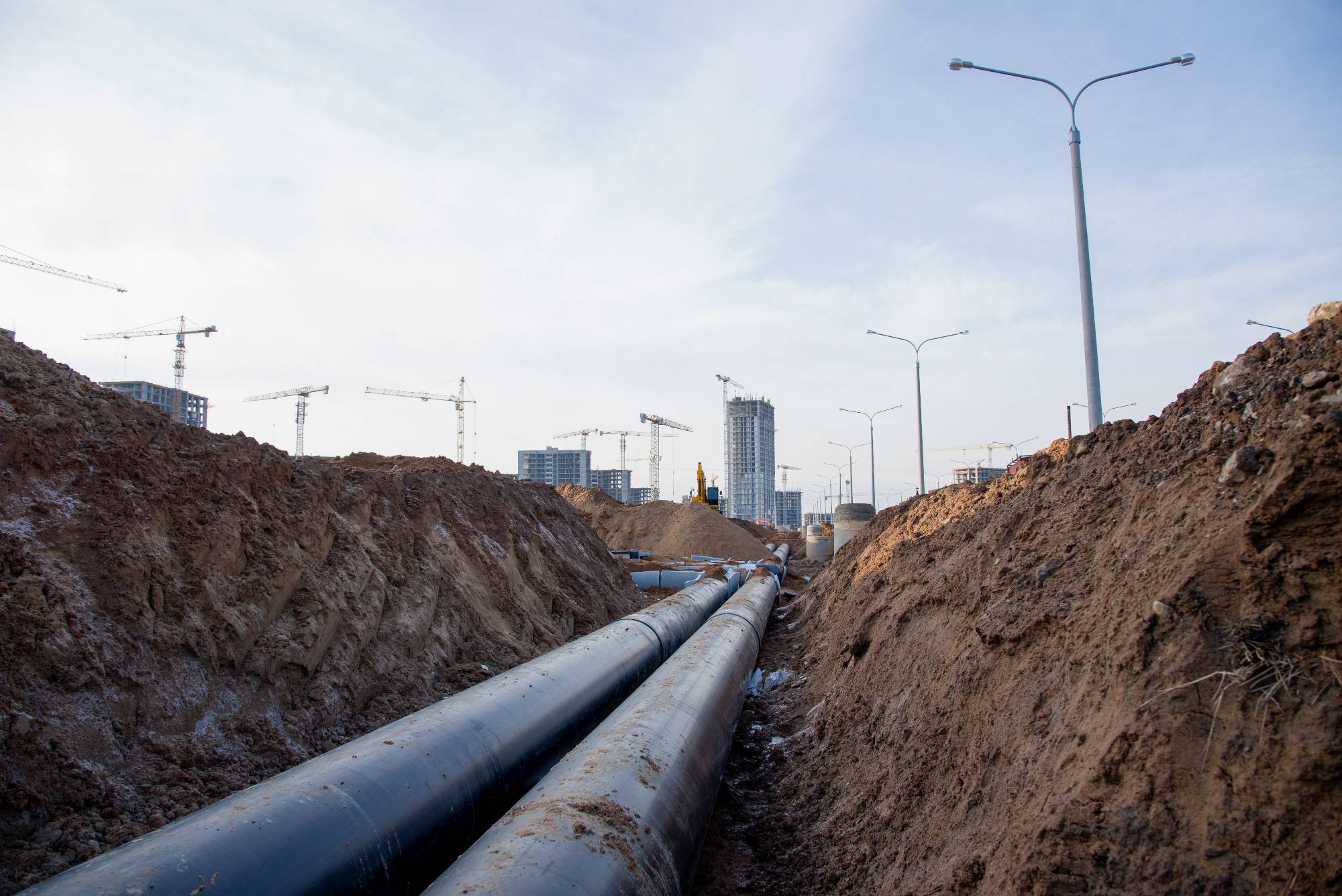 Large pipes run through a deep trench at a construction site, surrounded by dirt piles. Cranes and tall buildings rise in the background under a cloudy sky—another busy day for a plumber Bay Area, CA crews working along streetlights that line the road.