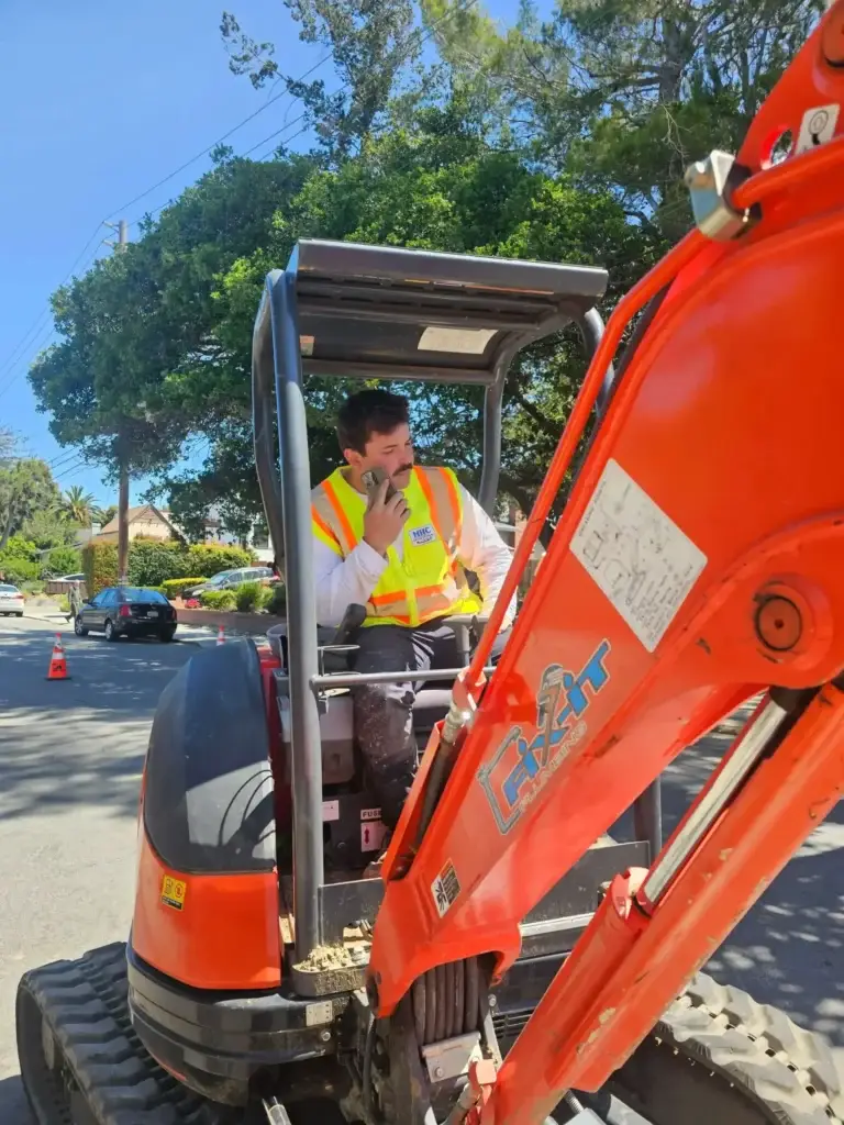 A plumber Bay Area, CA, wearing a white shirt and neon safety vest sits in the cab of an orange excavator on a sunny street, speaking into a handheld radio. Trees and parked cars are visible in the background.