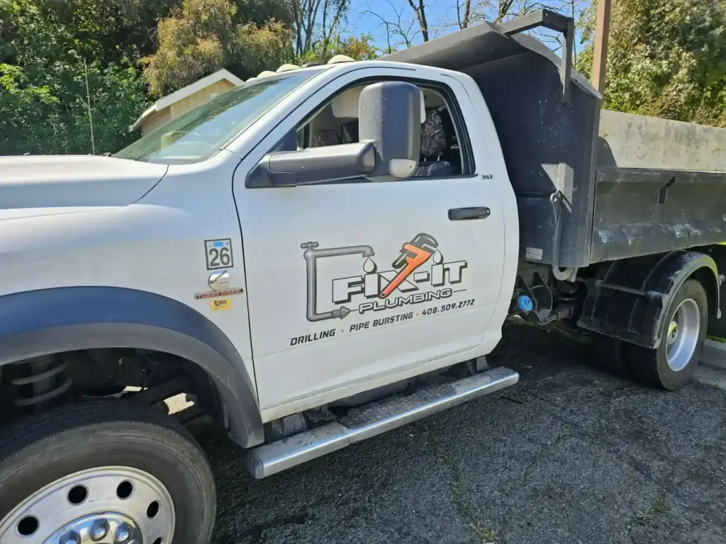 A white pickup truck with a Fixit Plumbing logo on the door, serving as a dump truck, is parked outside on a sunny day. This plumber Bay Area vehicle sits near trees and a house, ready for jobs across CA.