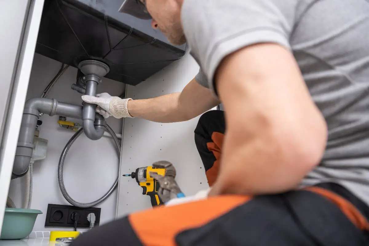 A plumber Bay Area professional wearing gloves and work clothes repairs pipes under a sink. Tools, including a drill and pliers, are nearby as the plumber works in a white cabinet space, focusing on the plumbing connections in CA.