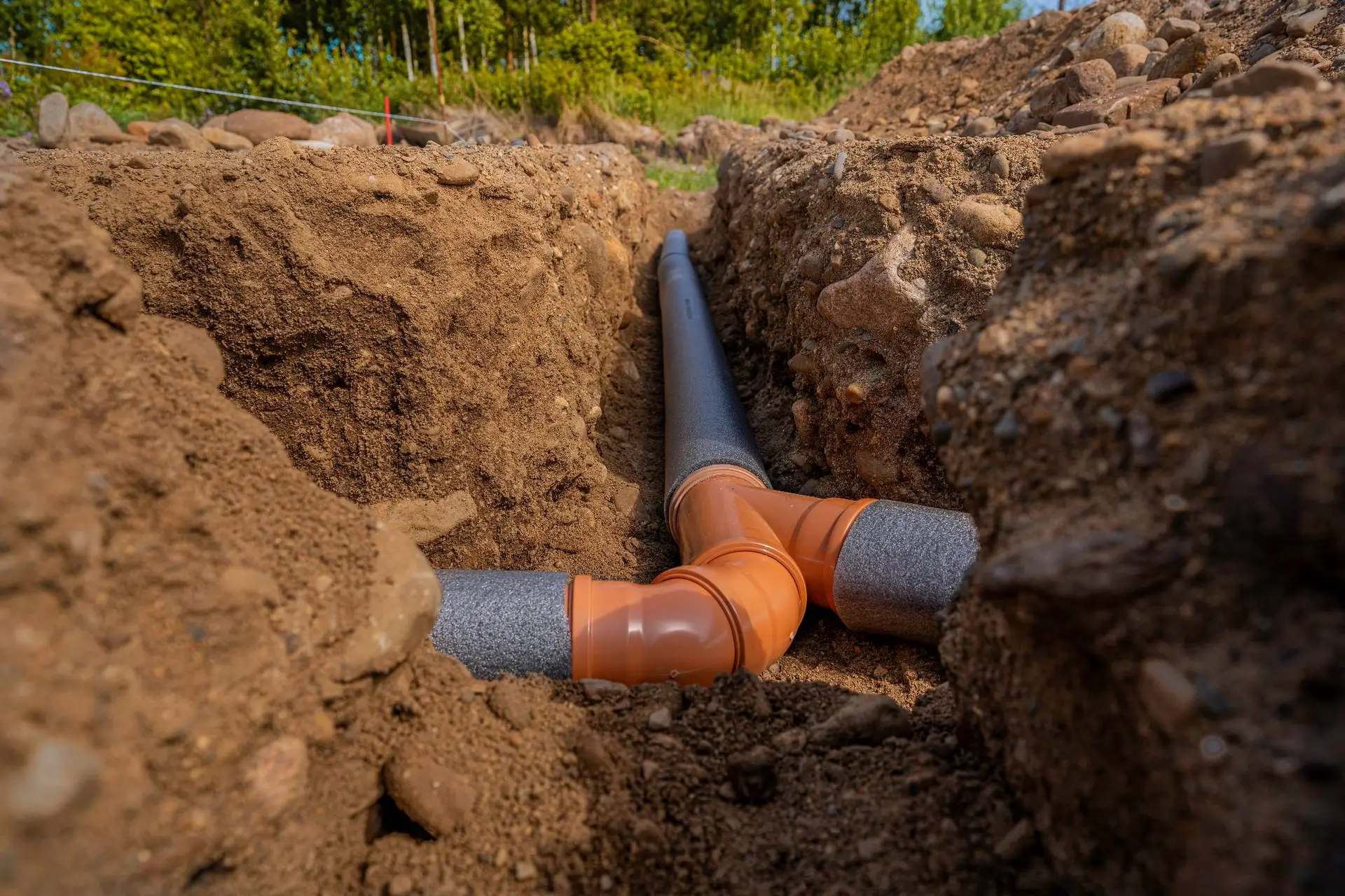 Two orange and gray plastic drainage pipes are connected underground in a dirt trench, surrounded by soil, with greenery in the background—typical work for a plumber Bay Area homeowners trust in CA.