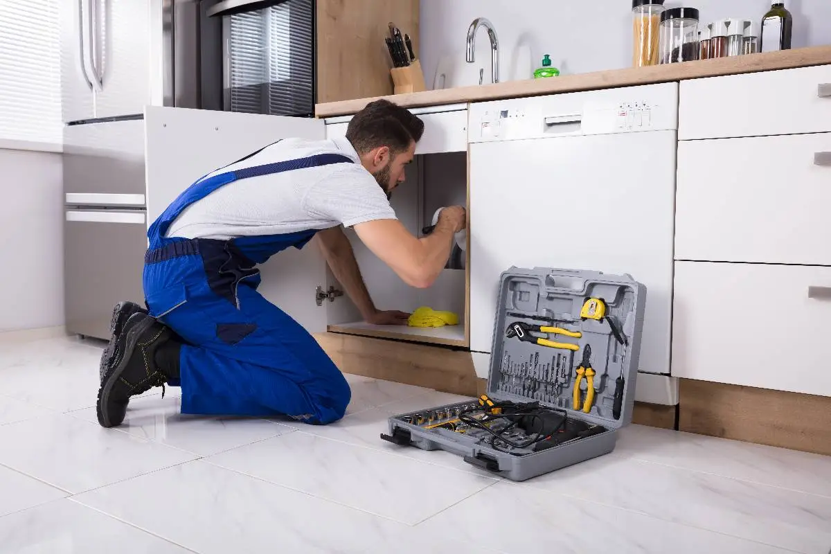 A plumber Bay Area in blue overalls kneels on a kitchen floor, fixing something under the sink. An open gray toolbox with tools is placed nearby. The kitchen has light cabinets and white tile flooring.