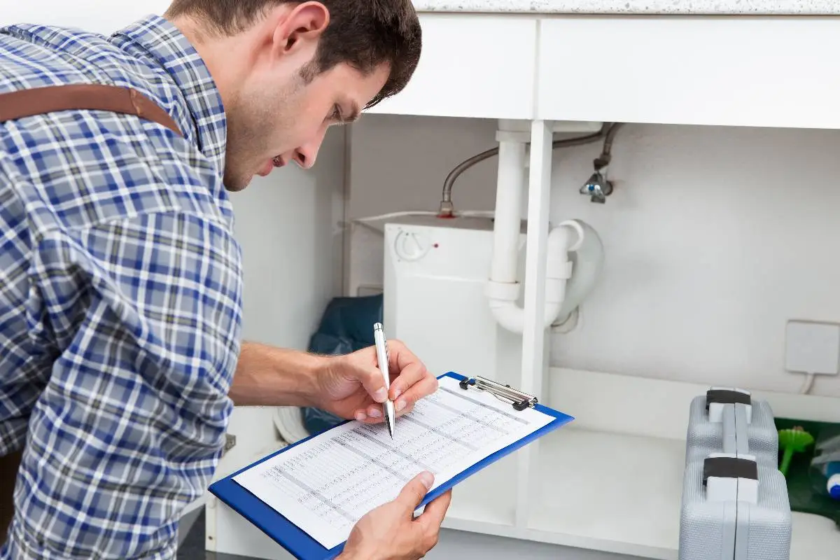 A plumber Bay Area professional in a plaid shirt fills out a checklist on a clipboard while inspecting plumbing under a sink. A toolbox is on the counter beside him, showing his dedication to quality service in CA.