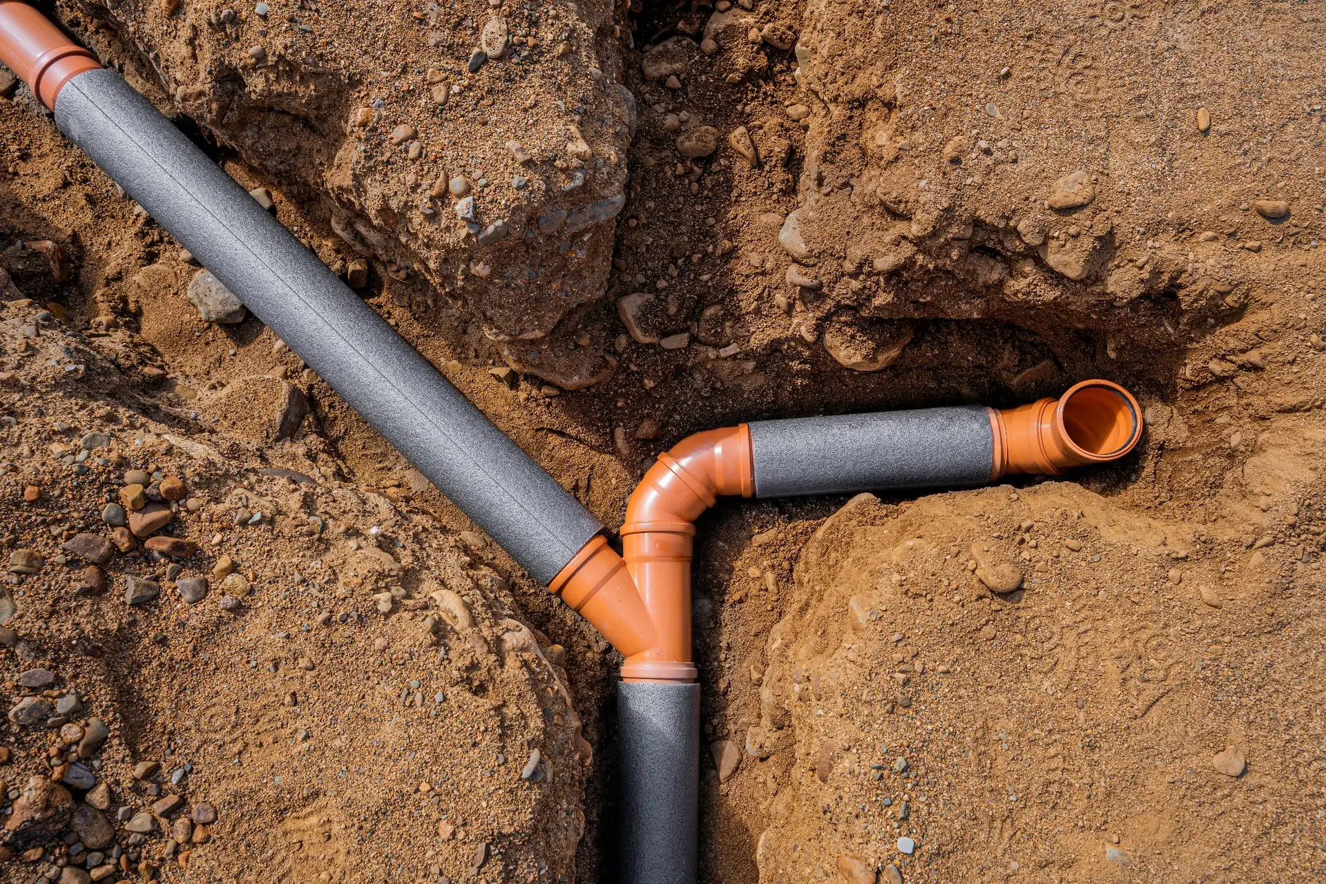 Two orange plastic pipes with black insulation are joined at an angle in a trench of brown soil, forming part of an underground plumbing system installed by a plumber Bay Area, CA.