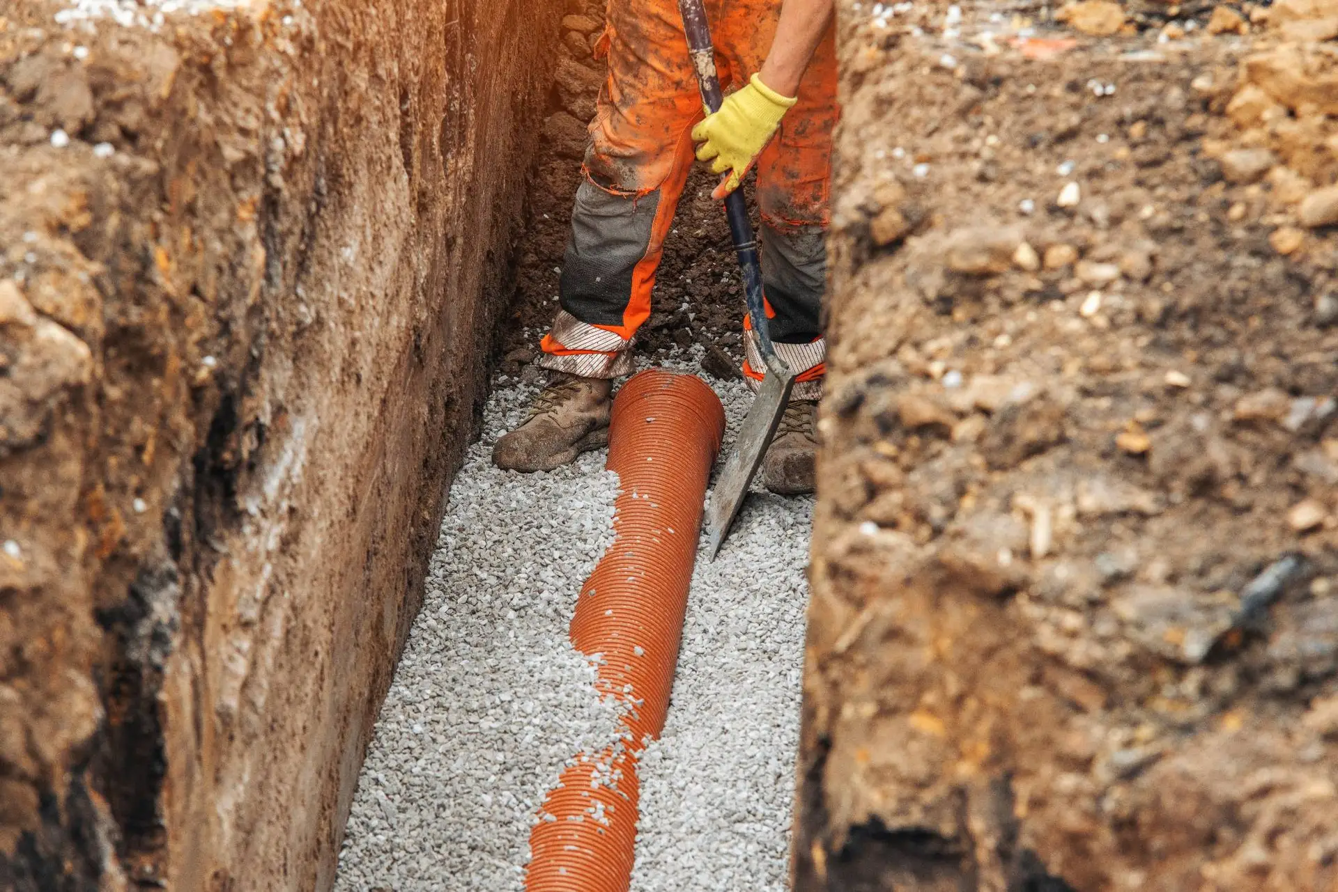 A plumber Bay Area, CA, in orange pants and yellow gloves uses a shovel to install a perforated orange drainage pipe in a gravel-filled trench at a construction site.