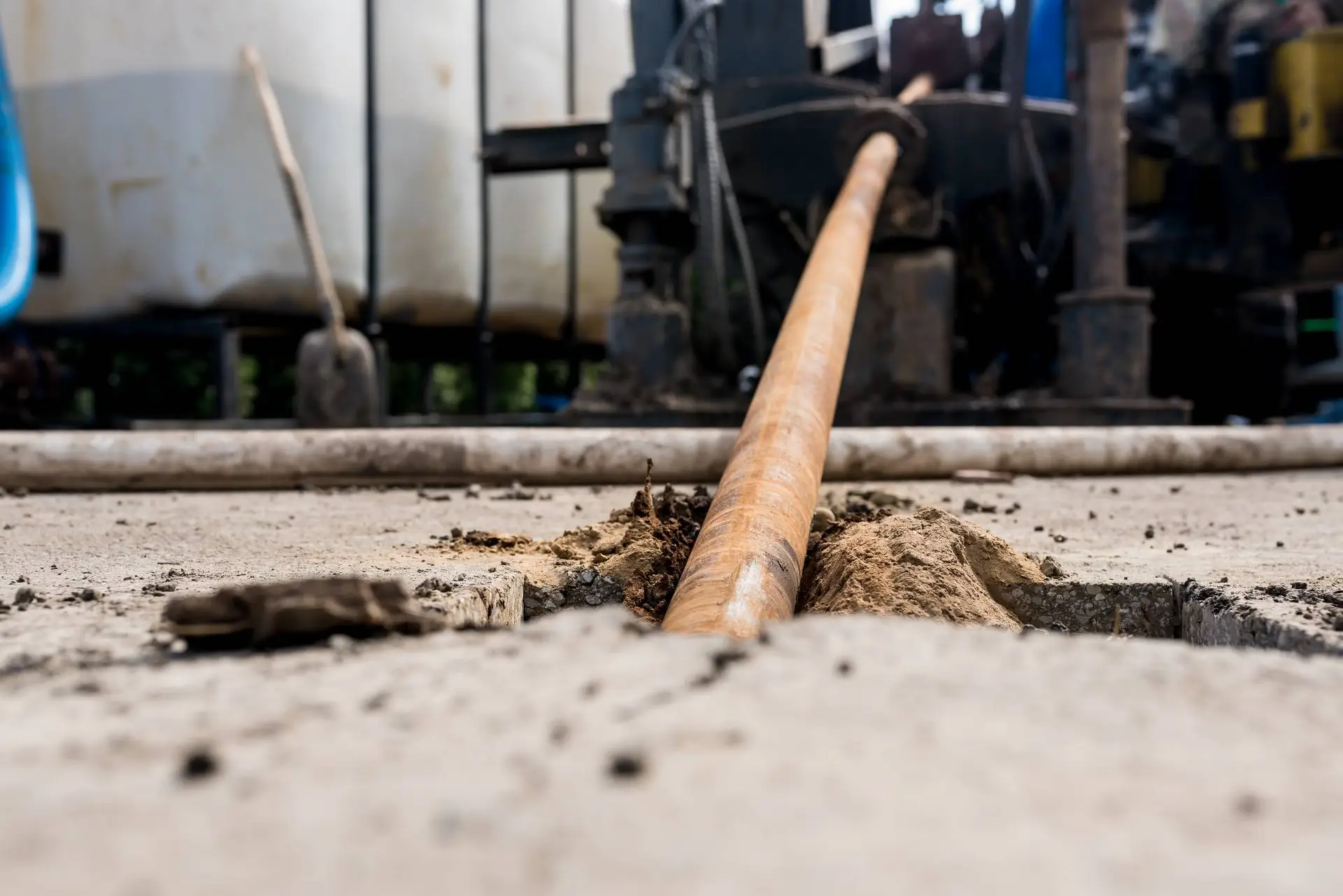 A large metal pipe is being drilled horizontally into the ground, displacing soil around the entry point; industrial equipment and tanks are visible in the blurred background, showcasing infrastructure work by a plumber Bay Area, CA.