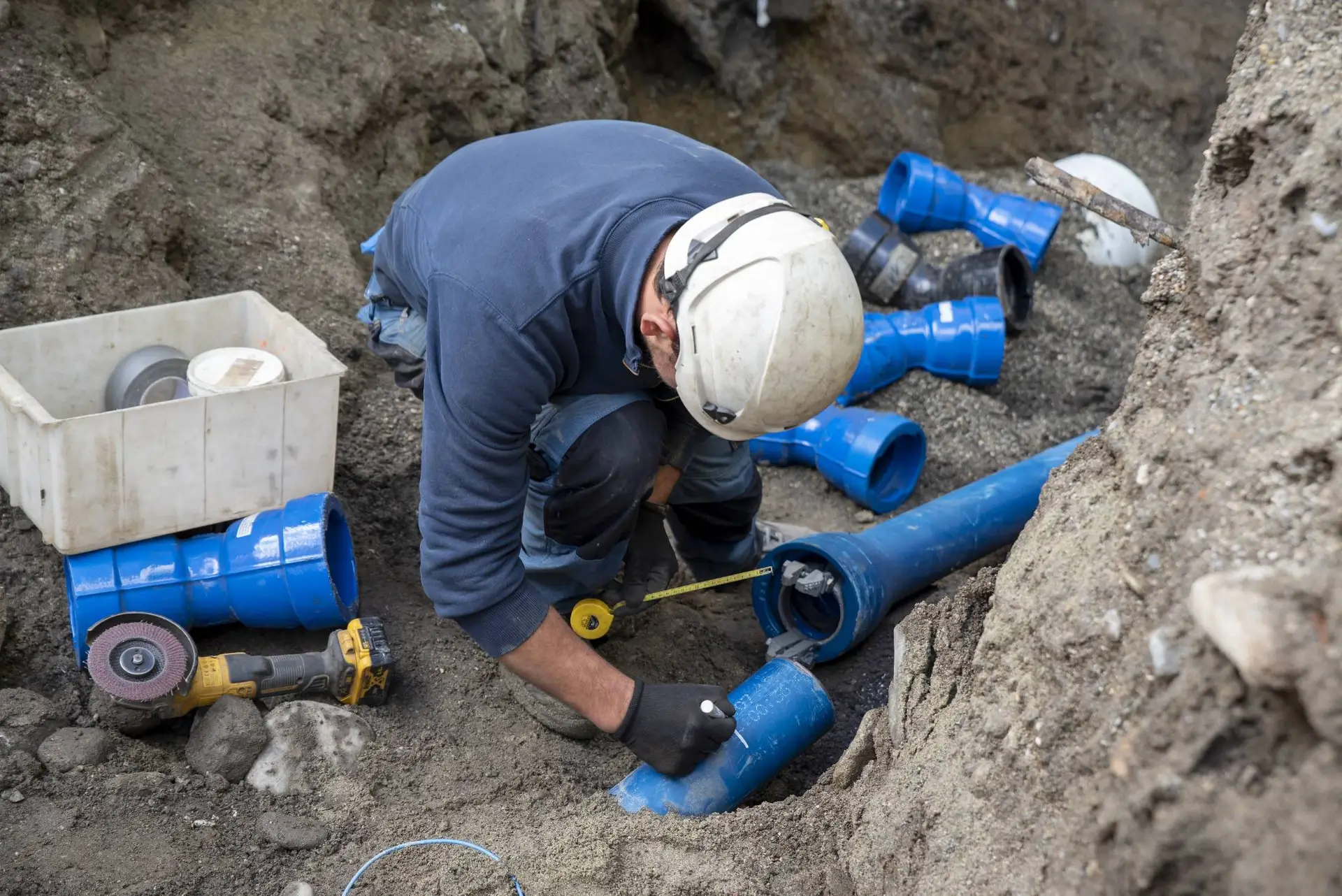 A construction worker wearing a white helmet and gloves measures and marks a blue plastic pipe at an outdoor excavation site in CA, surrounded by tools and pipe fittings—a typical day for a plumber Bay Area professionals.