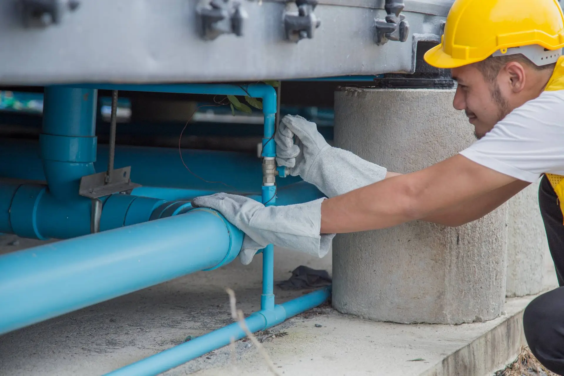 A plumber Bay Area worker wearing a yellow hard hat and gloves is inspecting or repairing blue pipes under a large structure outdoors in CA.