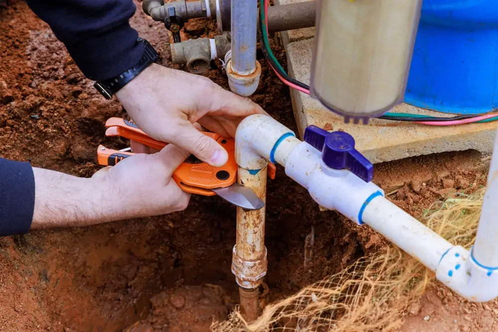 A plumber Bay Area, CA uses a pipe cutter to cut a white PVC water pipe connected to a rusty vertical metal pipe, with tools and plumbing fixtures scattered around the dirt area.