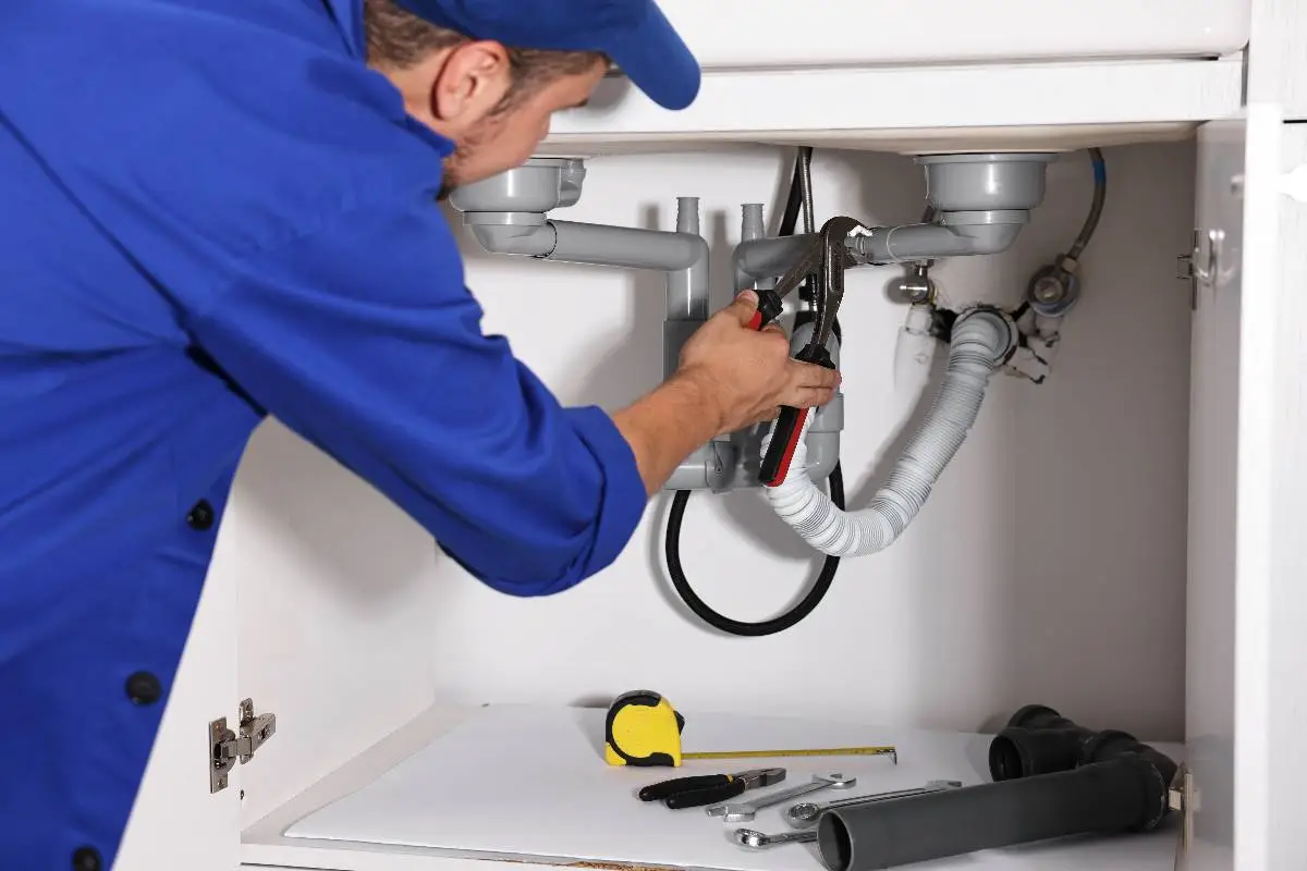 A plumber Bay Area in a blue uniform uses pliers to repair pipes under a sink. Tools and pipes are visible on the floor of the cabinet, showcasing expert service in CA.