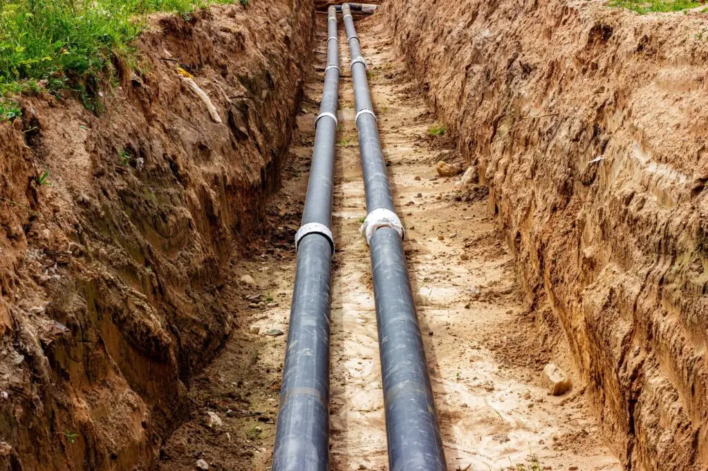 Two large black pipes are laid parallel in a long, narrow trench dug in the ground by a skilled plumber Bay Area, surrounded by dirt and soil, with some grass visible at the edges.