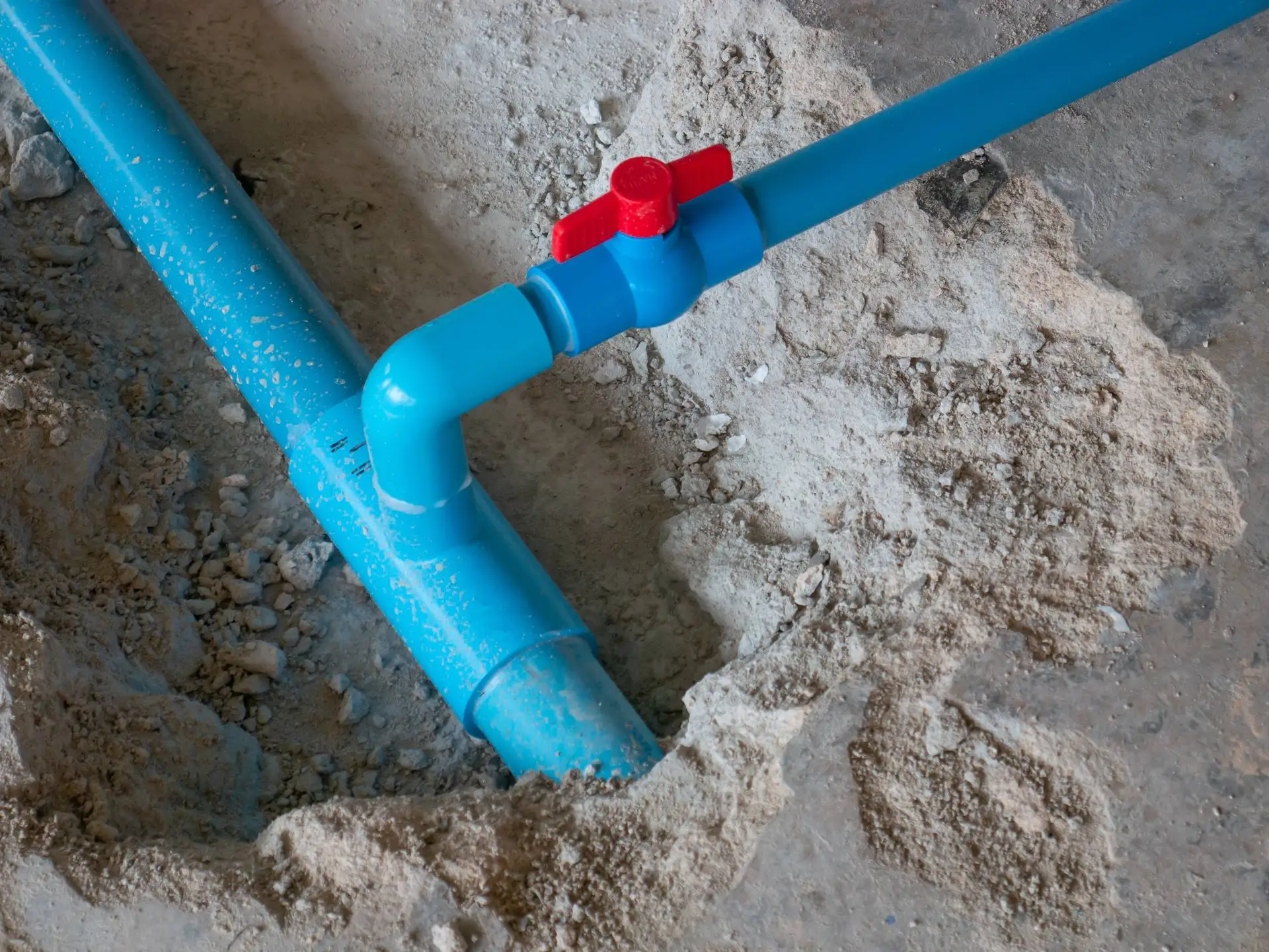 Blue plastic water pipes joined at a right angle with a red valve handle, partially buried in concrete or dirt—typical of installations by a skilled plumber Bay Area, CA.