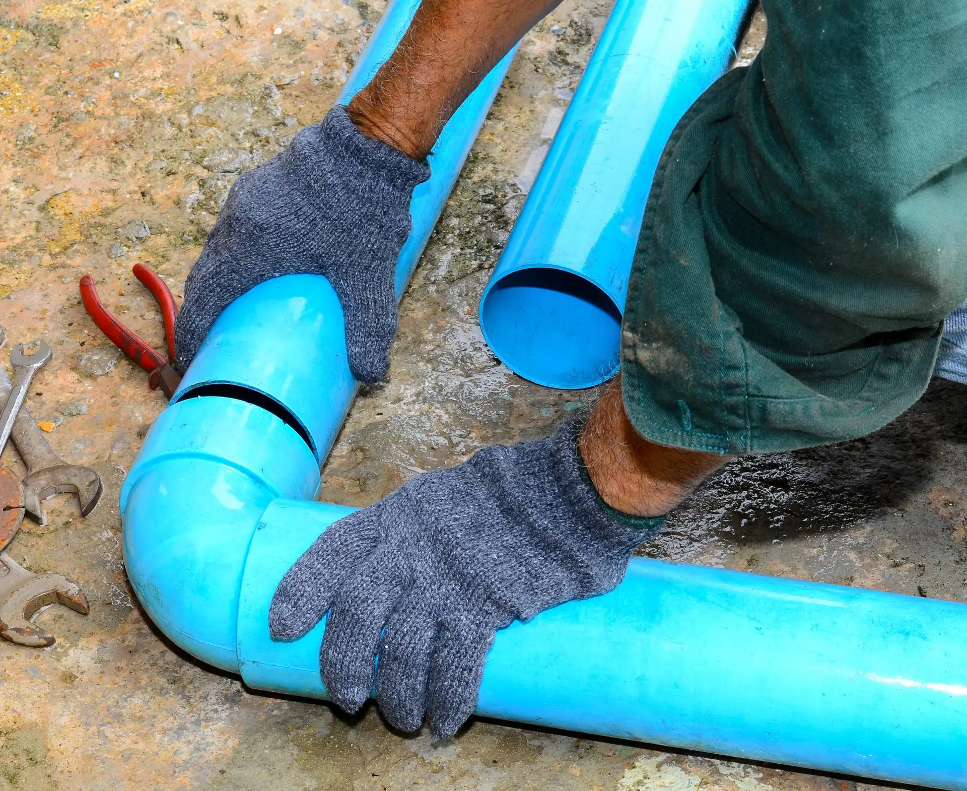 A plumber Bay Area in CA, wearing gray gloves and green sleeves, fits blue PVC pipes together on a concrete surface, with pliers and wrenches nearby.