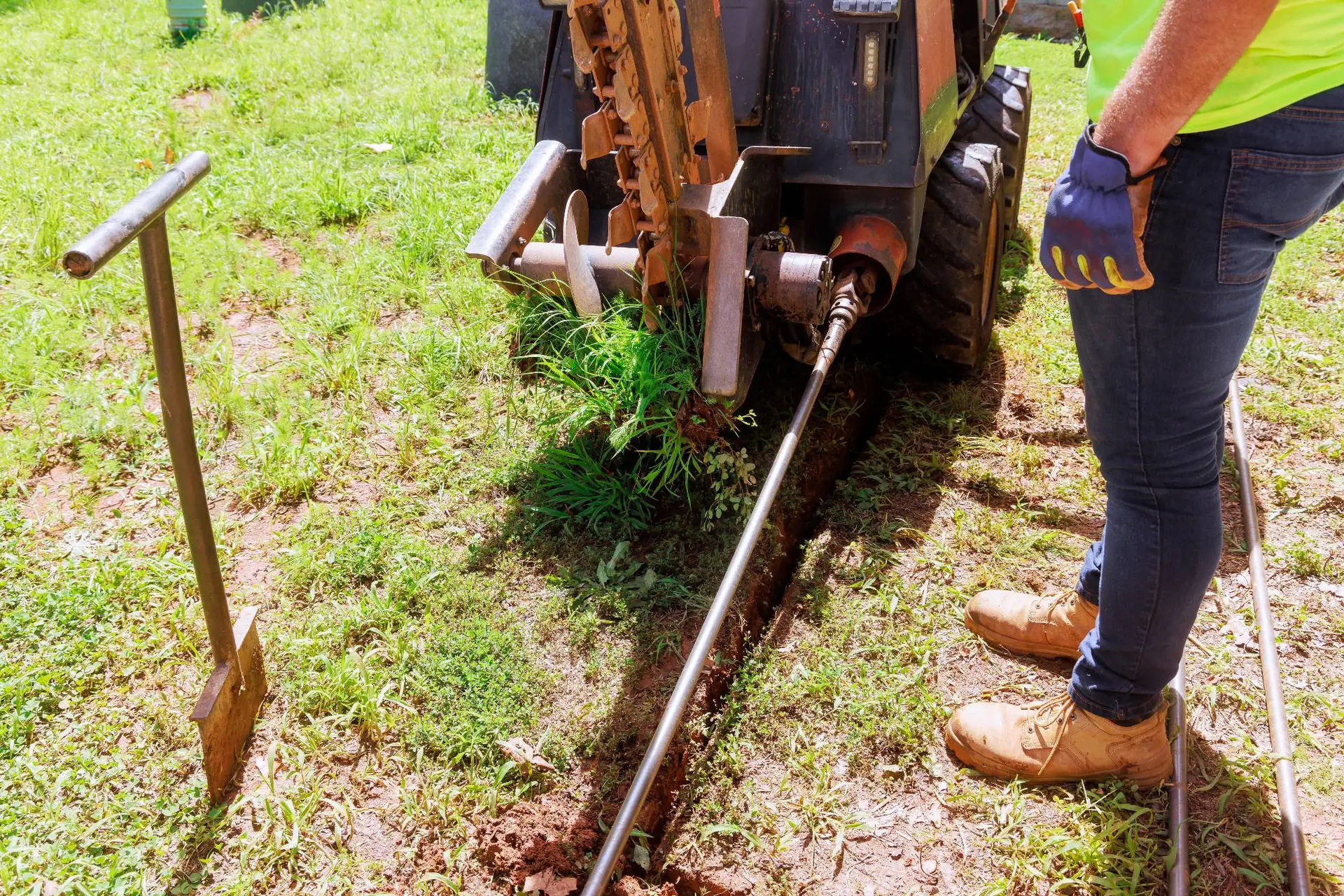 A plumber Bay Area in work gloves and boots operates a trencher machine, cutting a narrow trench in grassy CA ground. A shovel stands nearby, and the area is sunny with green grass.