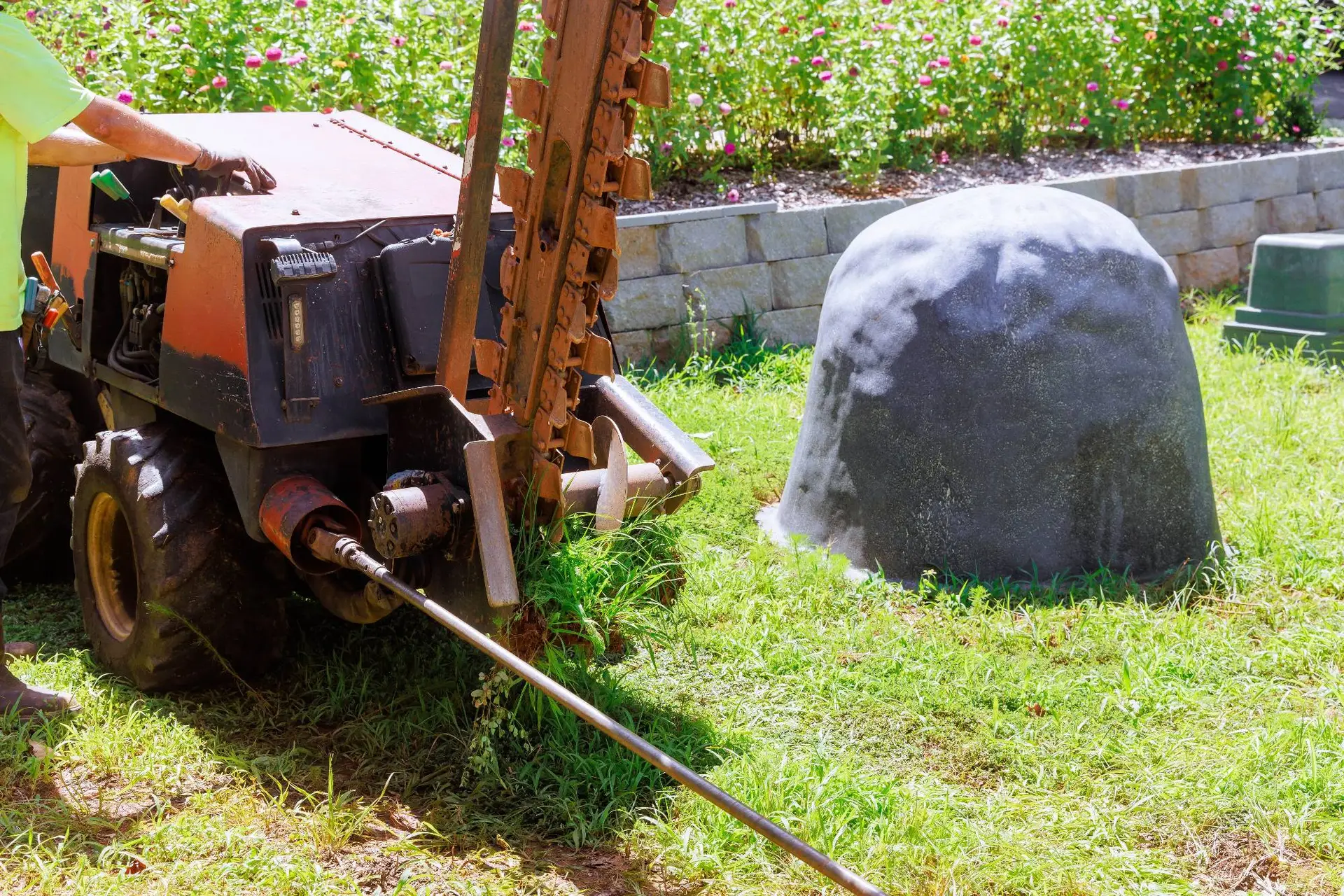 A plumber Bay Area worker operates a machine that drills or pushes a metal rod into the ground in a grassy CA yard, with a large gray rock or utility cover nearby and flowering plants in the background.